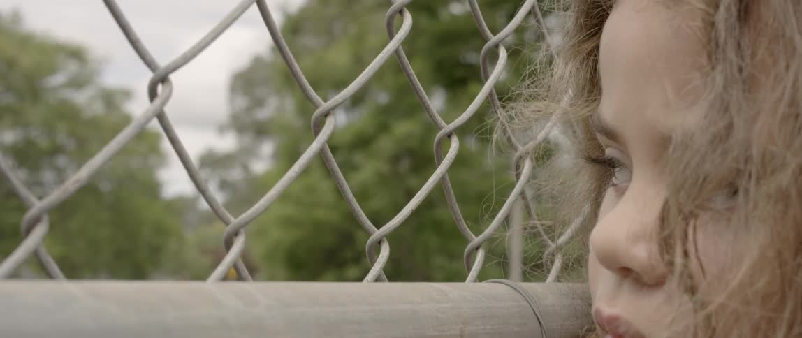 Child leans against a chain link fence looking sad