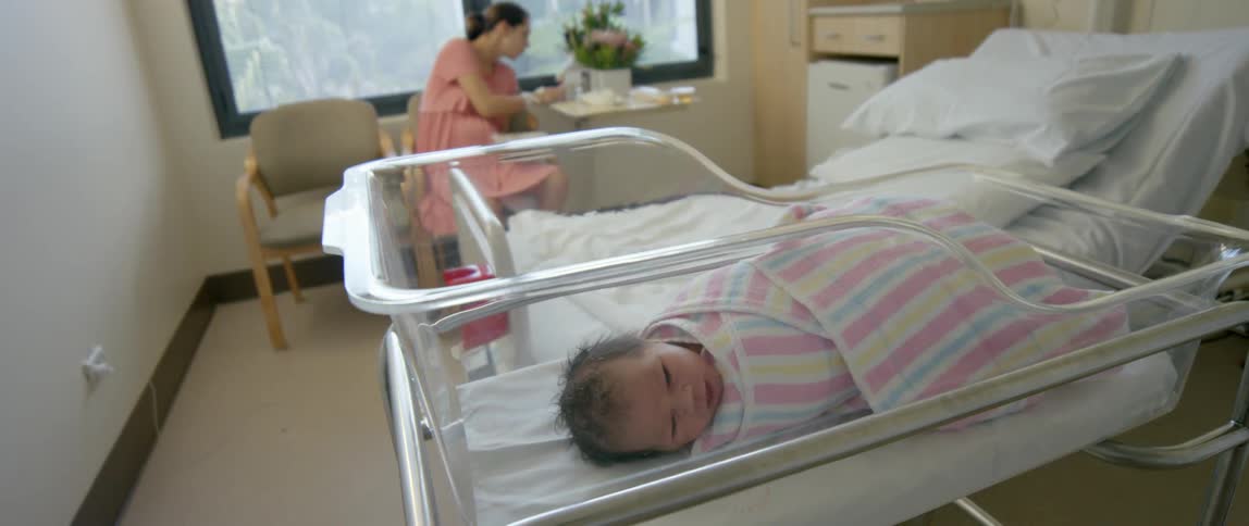 An infant lays in an incubator as a woman sits in a chair