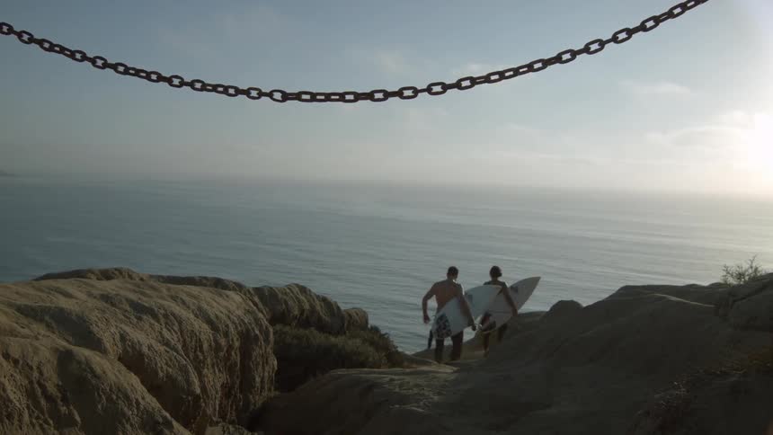 Men carrying surfboards as they climb down a rocky cliff towards the ocean
