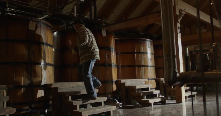 A distillery worker approaches a large vat with a graduated cylinder in hand