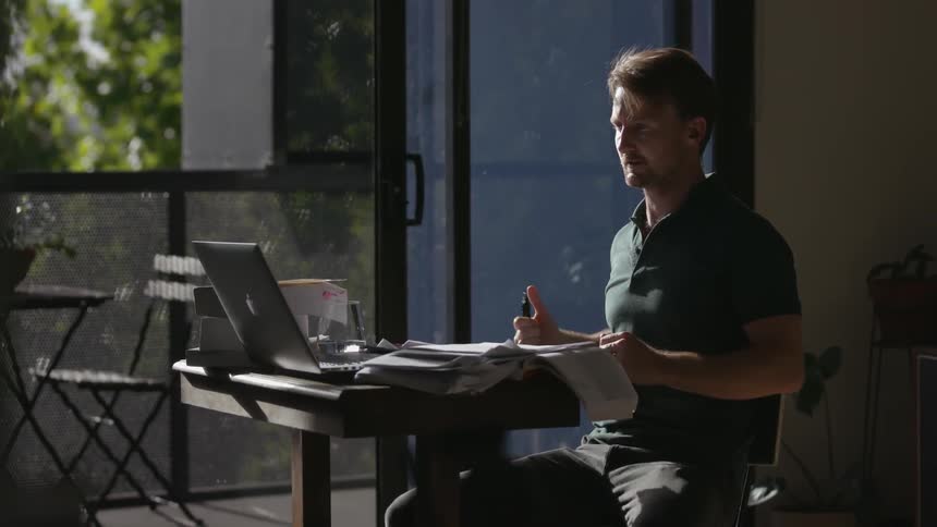 A man sits down at his desk near the window with a pen