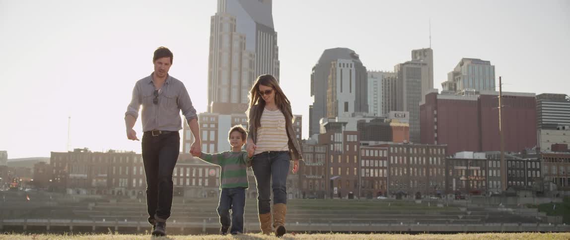 A man, woman and child are holding hands walking through a field with a cityscape in the background