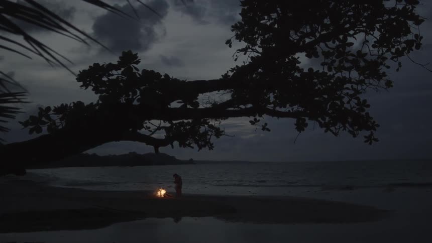 A couple stand by a bonfire on a beach at dusk