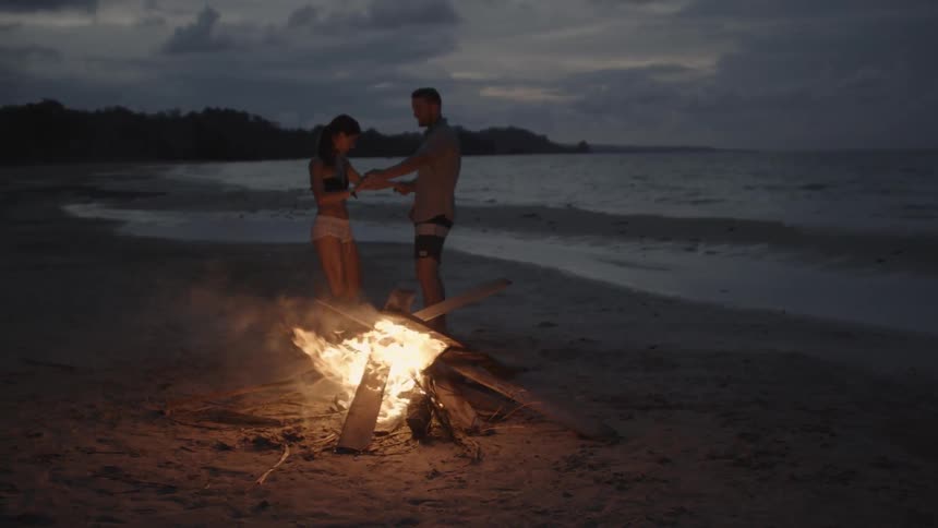 A man and woman dance by a bonfire on a beach