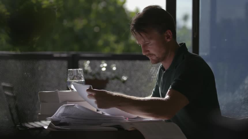 A man sits at a small table while looking through a stack of papers