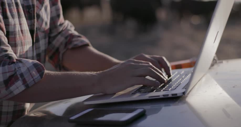 A man types on a laptop sitting on a truck hood