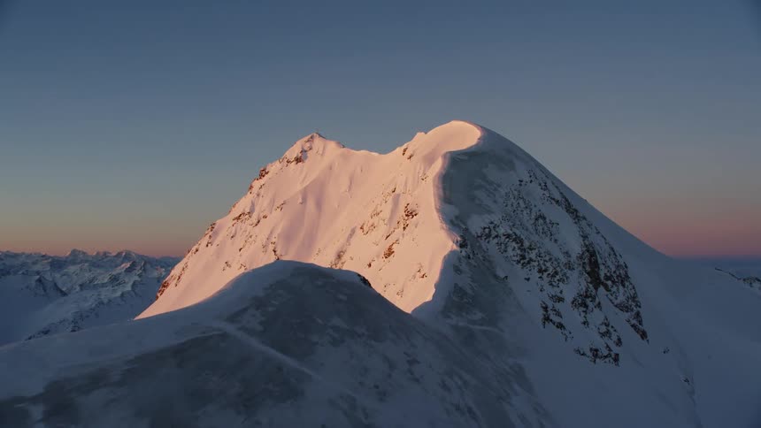 The sun casts a pink glow on a snow-covered mountain peak