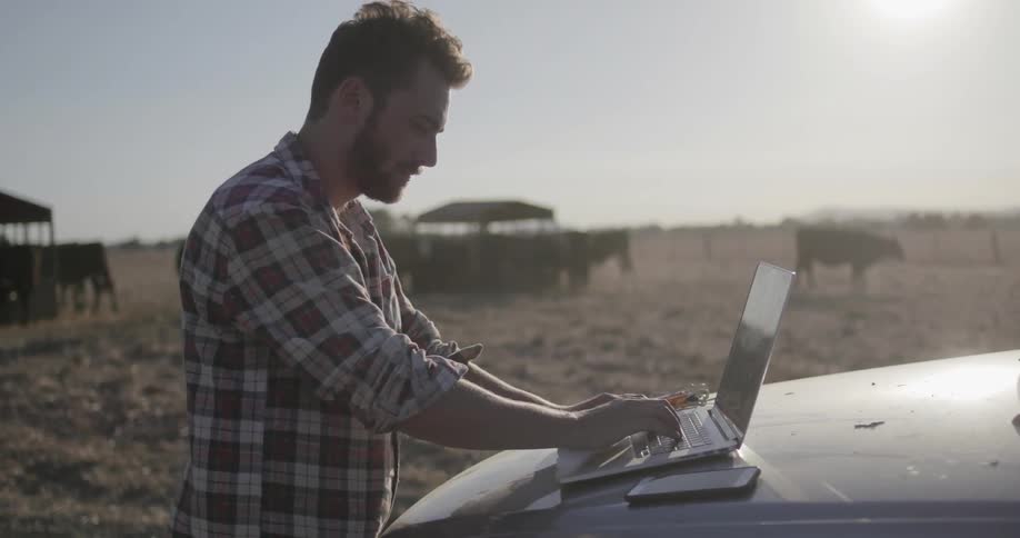 A man on a farm types on his laptop which is on a car hood