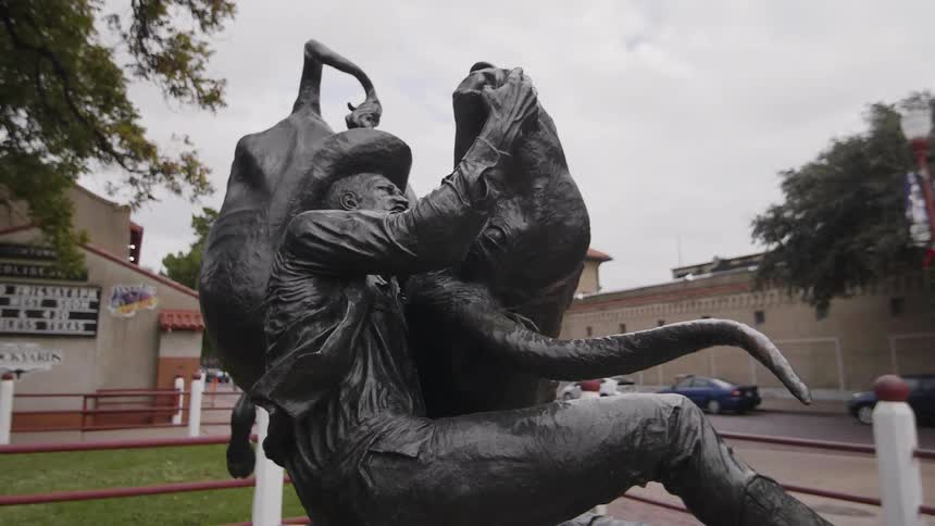 A metal statue of a man wrestling a longhorn bull sits in a park like setting