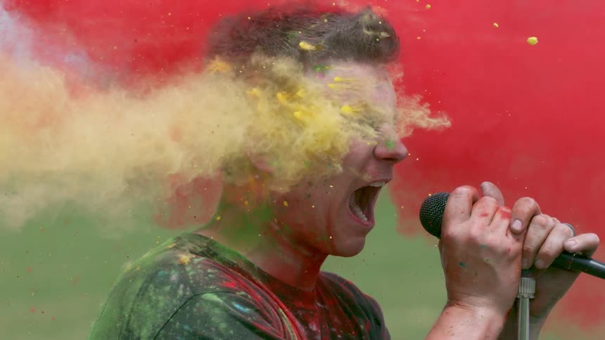 A man screams into a microphone as his face is struck with colorful powder