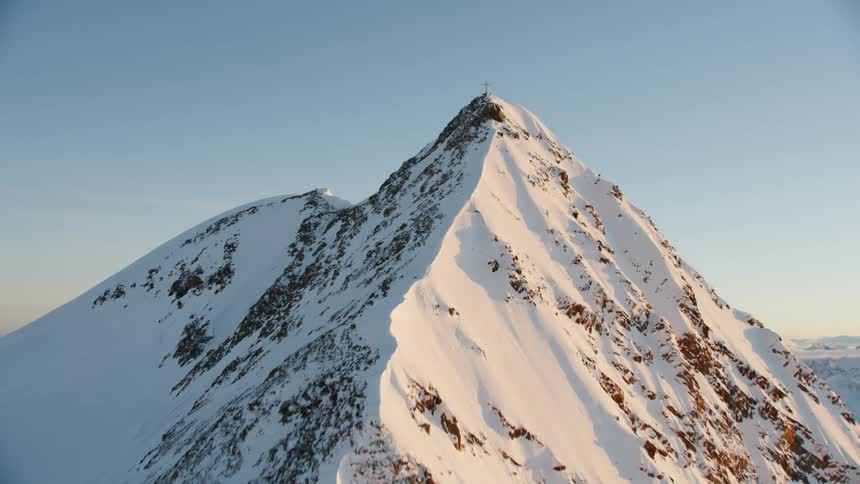 A cross stands high atop a peak in a snow-covered mountain range