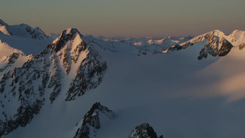 The sun casts a pink glow on a snow-covered mountain range