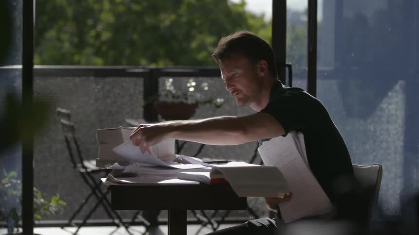 A man sitting at a table near a balcony looks through piles of paper