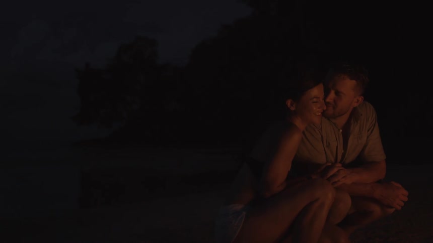 A man kisses a woman as they sit near a fire on the beach