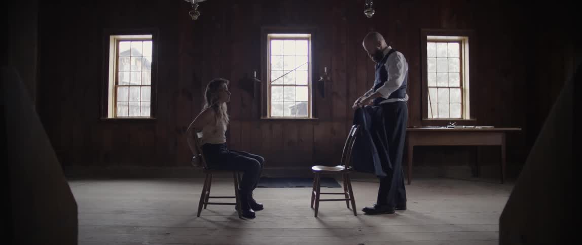 A man brings a chair over to a woman who is handcuffed to another chair