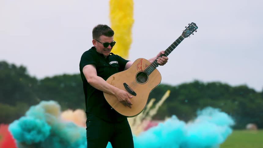 Man plays a guitar in a field and different colors of powder explode behind