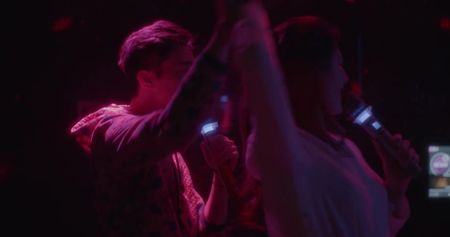A man and woman sing karaoke in a dark room with rotating lights