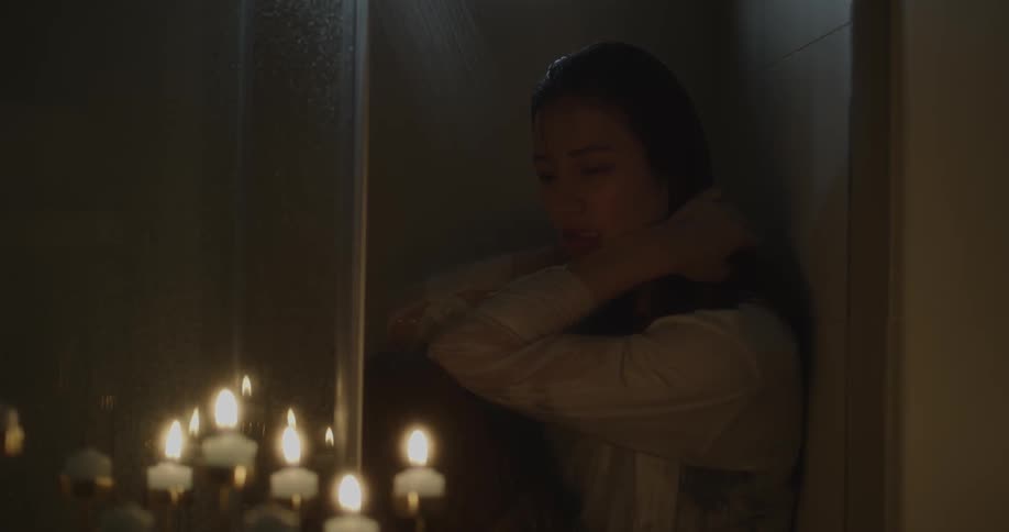 A woman sits in a running shower with her clothes on and candles flickering nearby