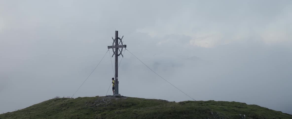 A man stands beside a cross on a grassy peak