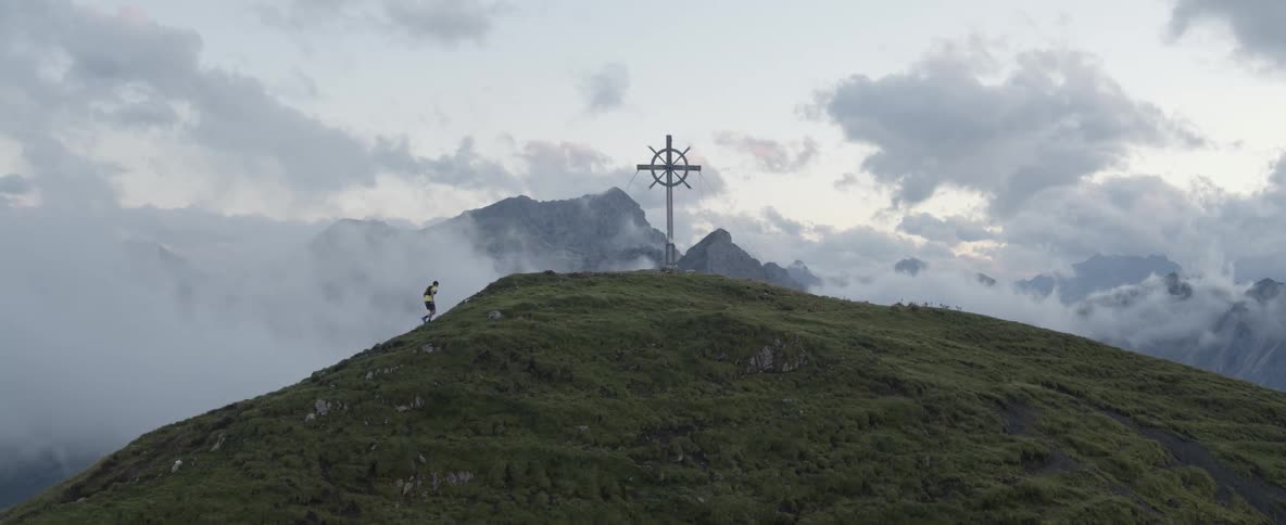 Man jogs up a grassy mountain on a cloudy day towards a cross