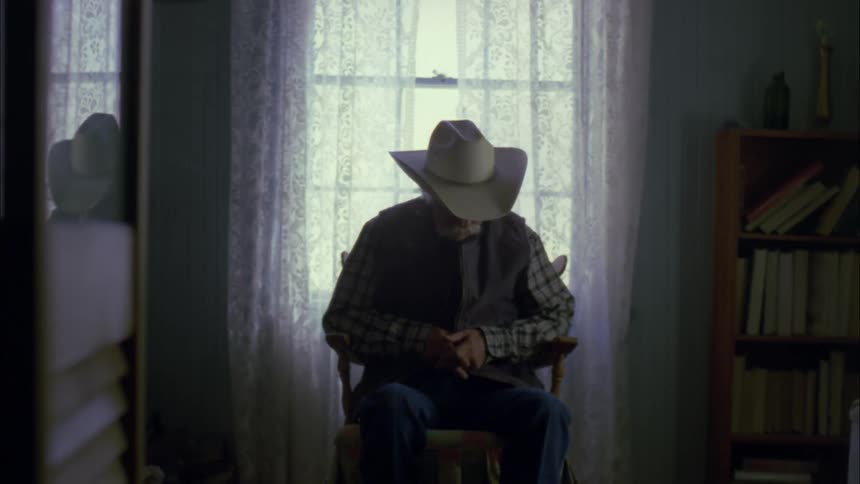 Older man in cowboy hat sitting in chair in house next to window