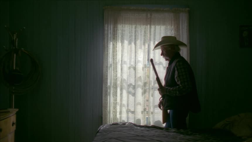 An old man sitting on a bed rises and retrieves his rifle