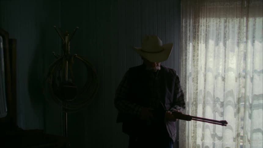 An old man gets up from sitting on a bed and cocks the gun he is holding