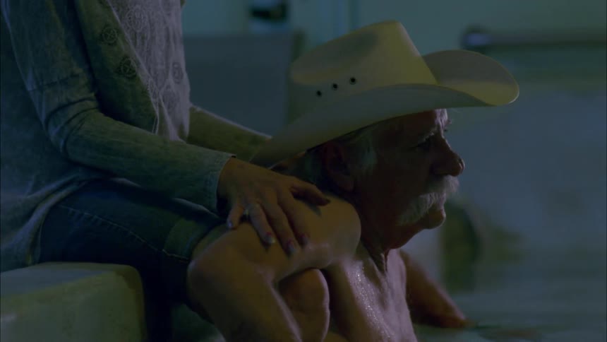 An elderly cowboy sits in a spa bath while having his shoulders rubbed