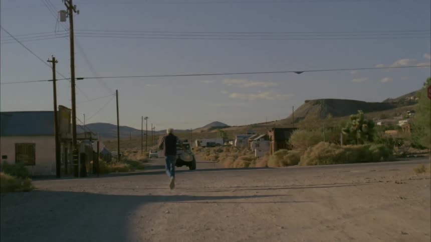 Man runs down a gravel road waving his hat at a vehicle
