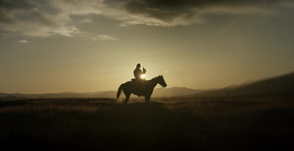 Man sitting on horse puts cowboy hat on at sunset
