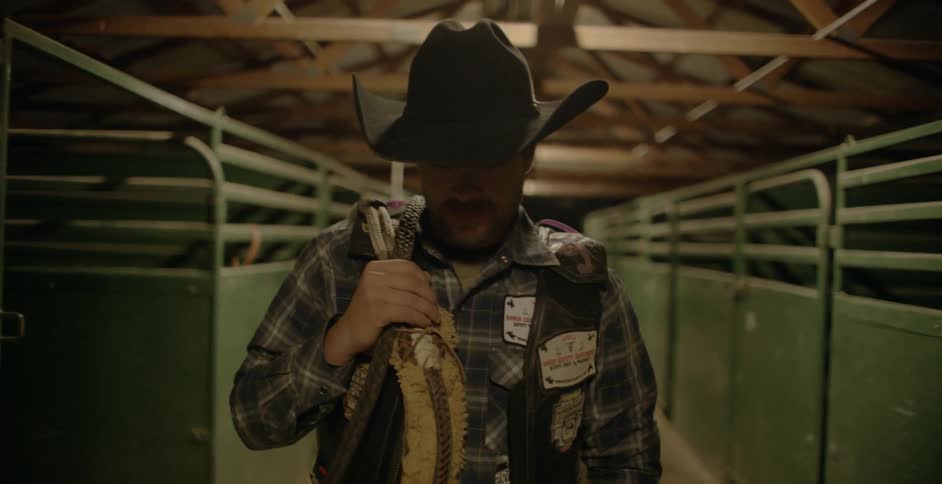 A cowboy carrying a strap on his shoulder walks between horse stalls