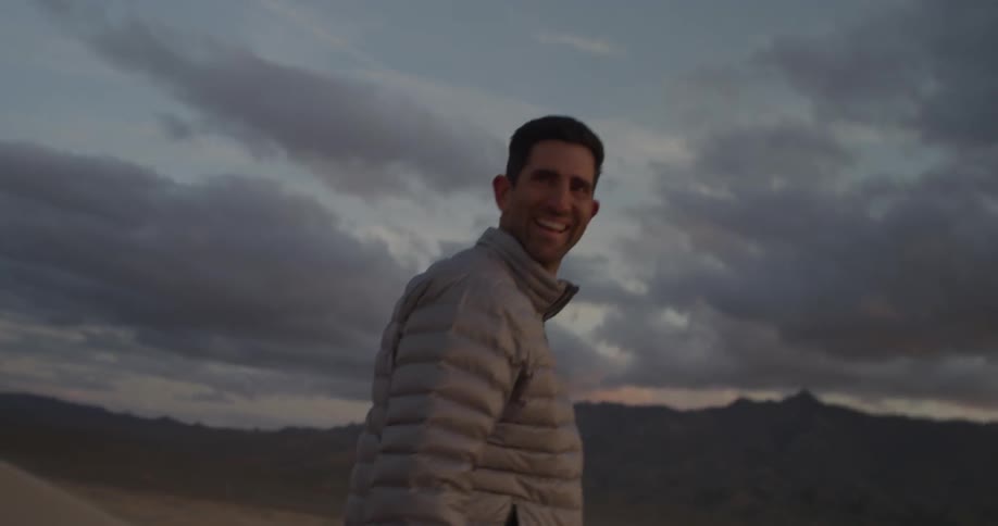A man smiles while walking near a mountain at sunset