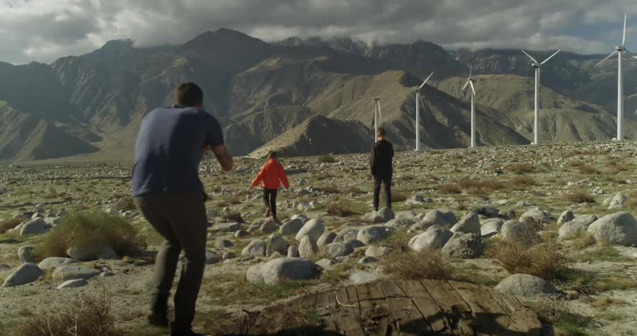 A man films two boys and a dog walking across a rocky field