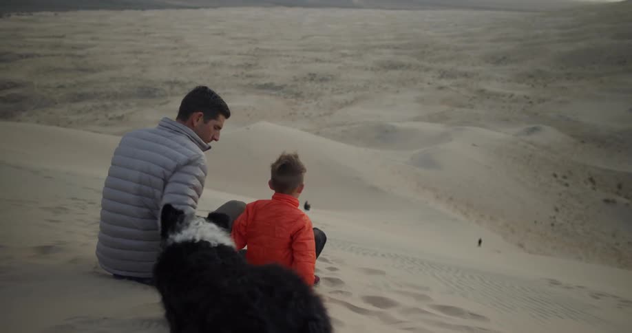 A man sits with a boy and dog on a sand dune in the desert