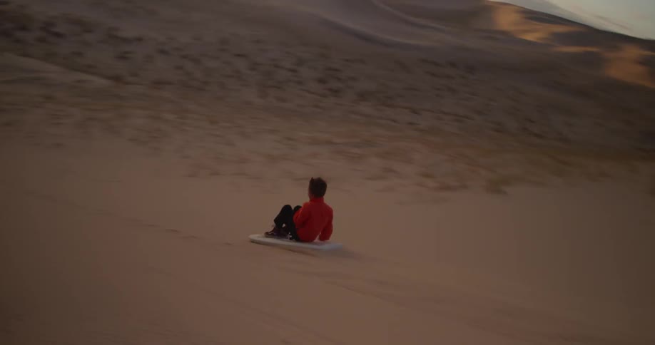 A boy rides a board down a sand dune