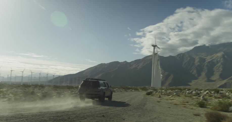 A man drives an suv down a dirt road towards windmills