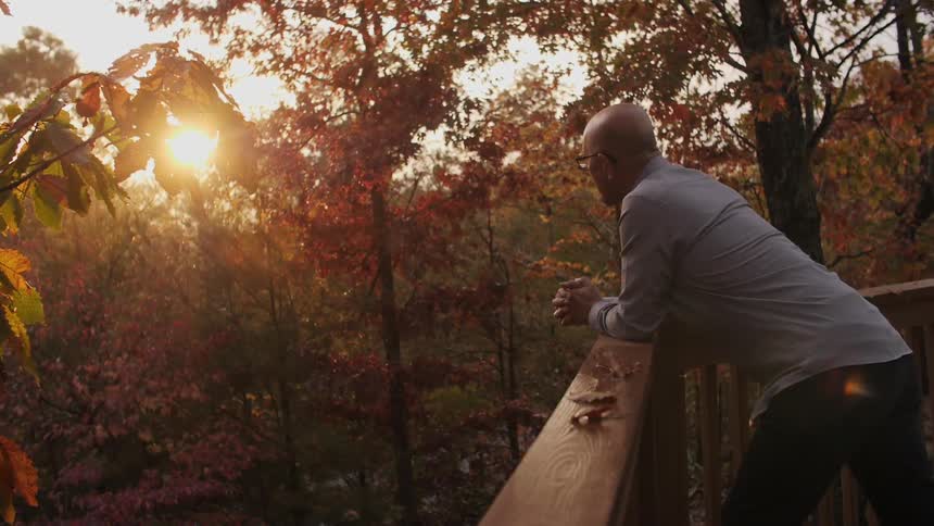 A man stands on a deck overlooking fall trees at sunset