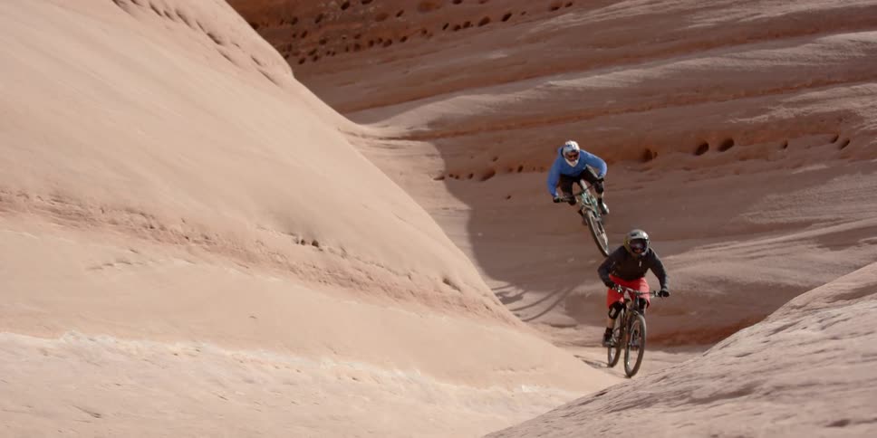 Two people ride their bikes over sandstone formations on a sunny day