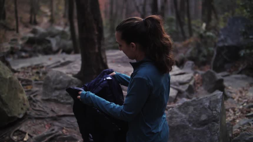 A woman unfolds a jacket as she rises to stand in a forest during the day