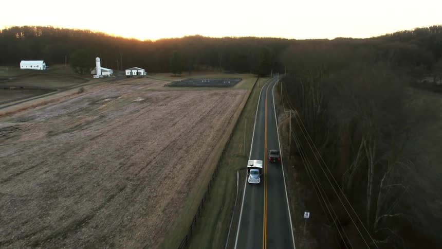 A vehicle towing a trailer drives past a farm on a country road