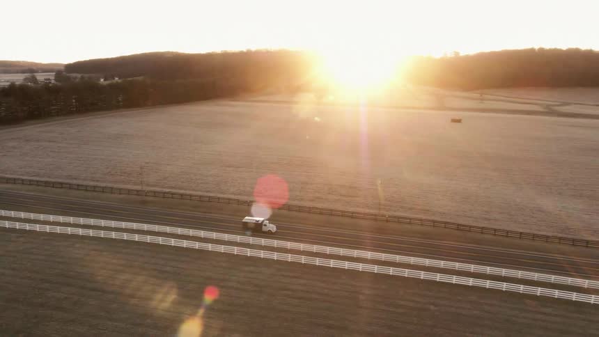 At sunrise, a truck drives along a road surrounded by open fields