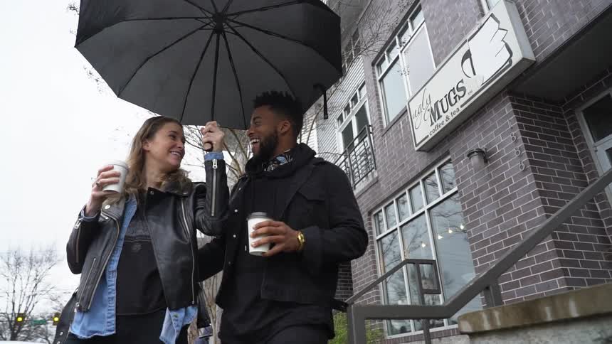 A man and woman hold coffee cups and walk under an umbrella