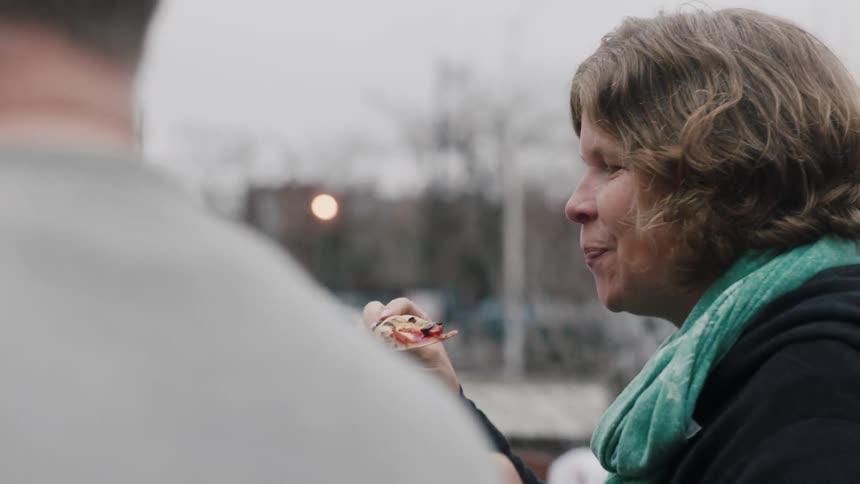 A woman eats outside and converses with a man