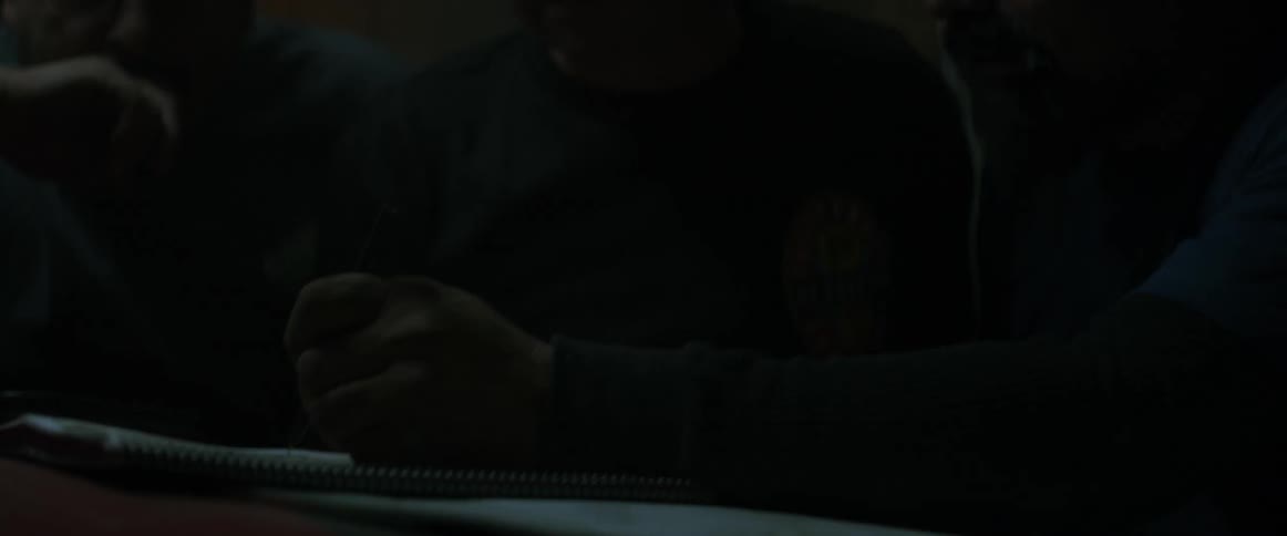 A man jots down notes in a notebook and converse with two others