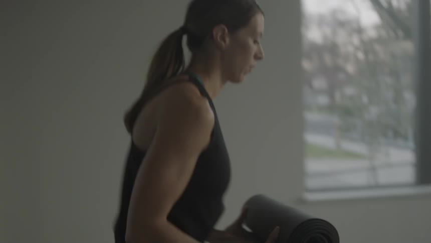 A woman carries a yoga mat into a room and unrolls it