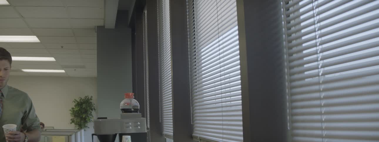 A man holding a cup looks through the blinds at work