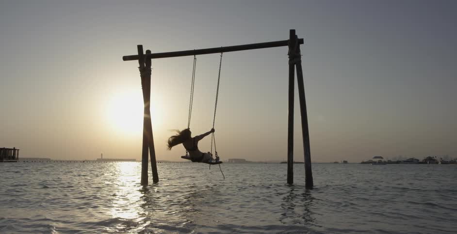 A woman is swinging on a swing set over the water at a beach