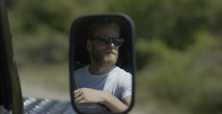 A man sitting in an suv on a sunny day is reflected in the passenger side mirror