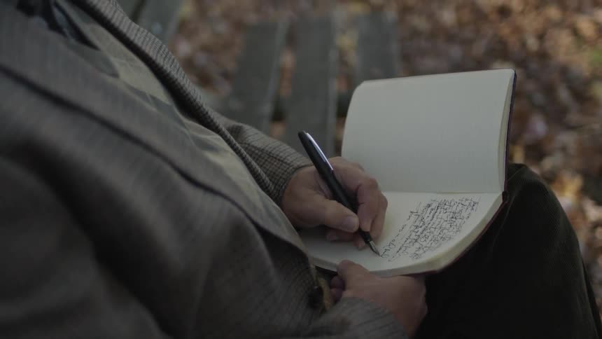 A man writes in a notebook while sitting on a park bench