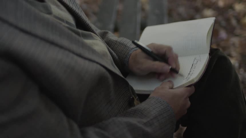 Person writes in a small book outside on a bench